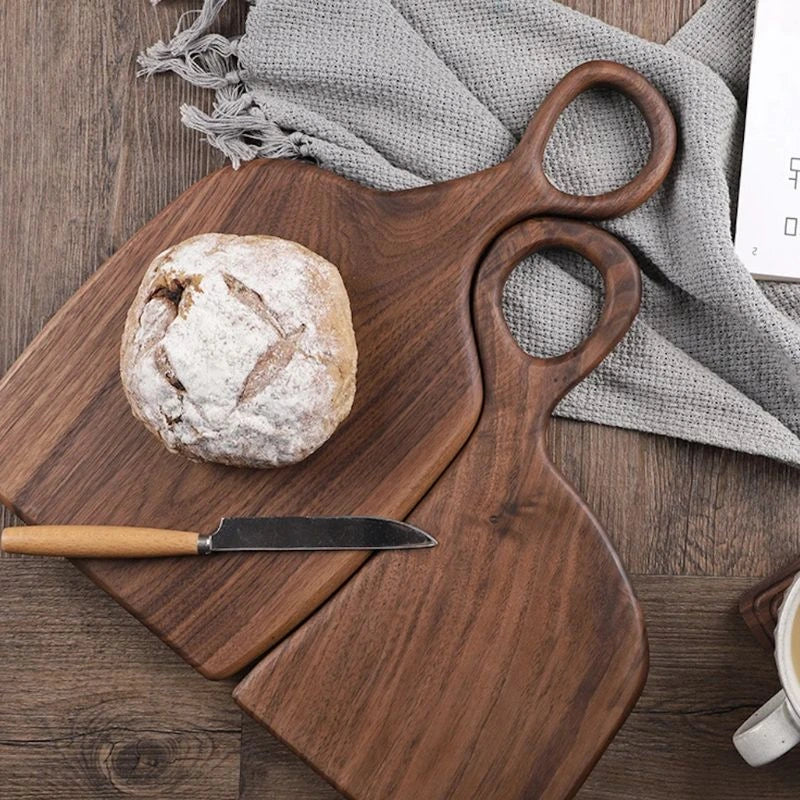 Zwei Schneidebretter aus dunklem Holz mit Brot und Messer auf einem Tisch mit grauem Tuch.