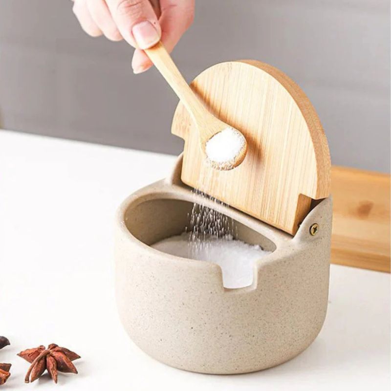 Hand hält Holzlöffel über geöffneter Keramikdose mit Gewürz.